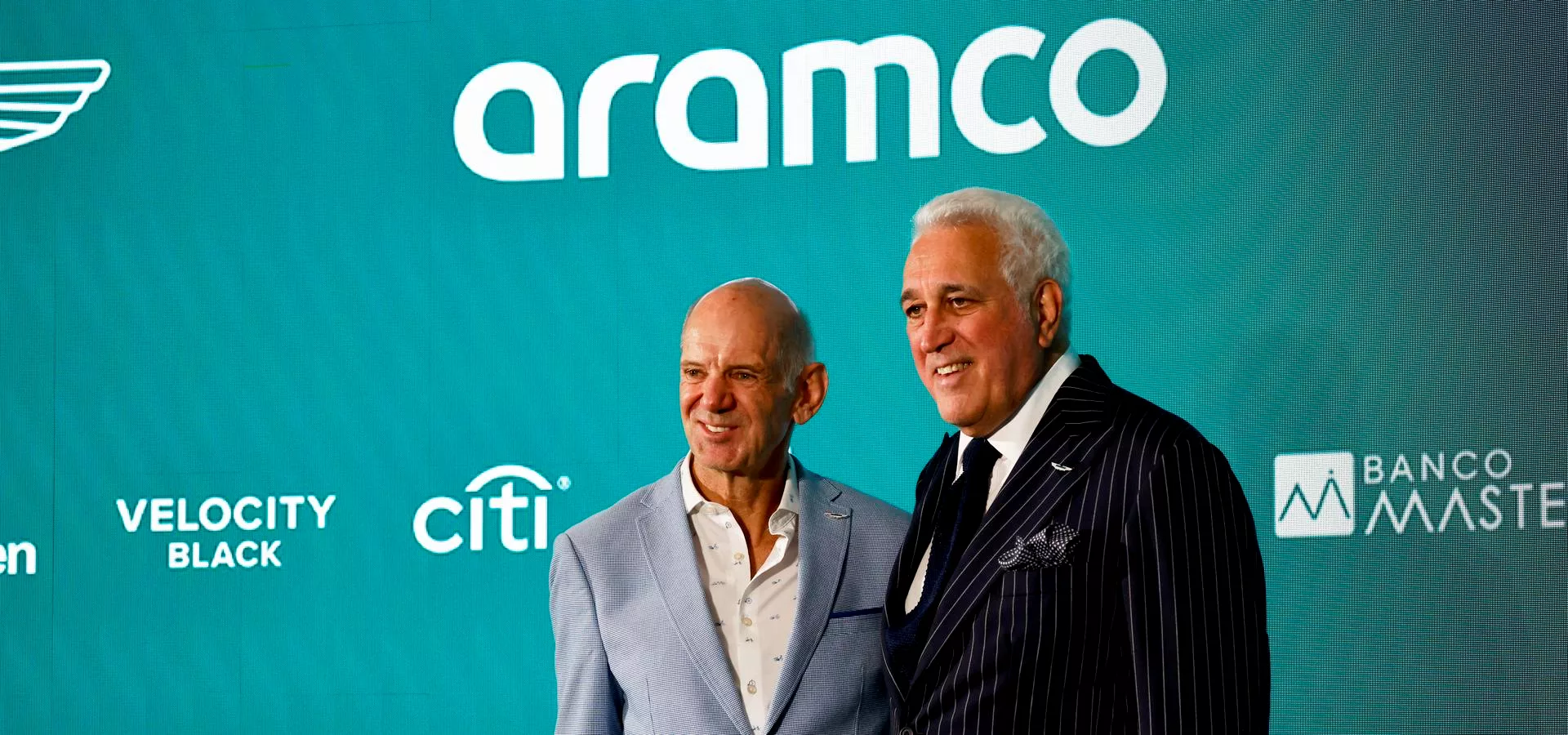 Two men wearing suits, smiling, in front of a green background
