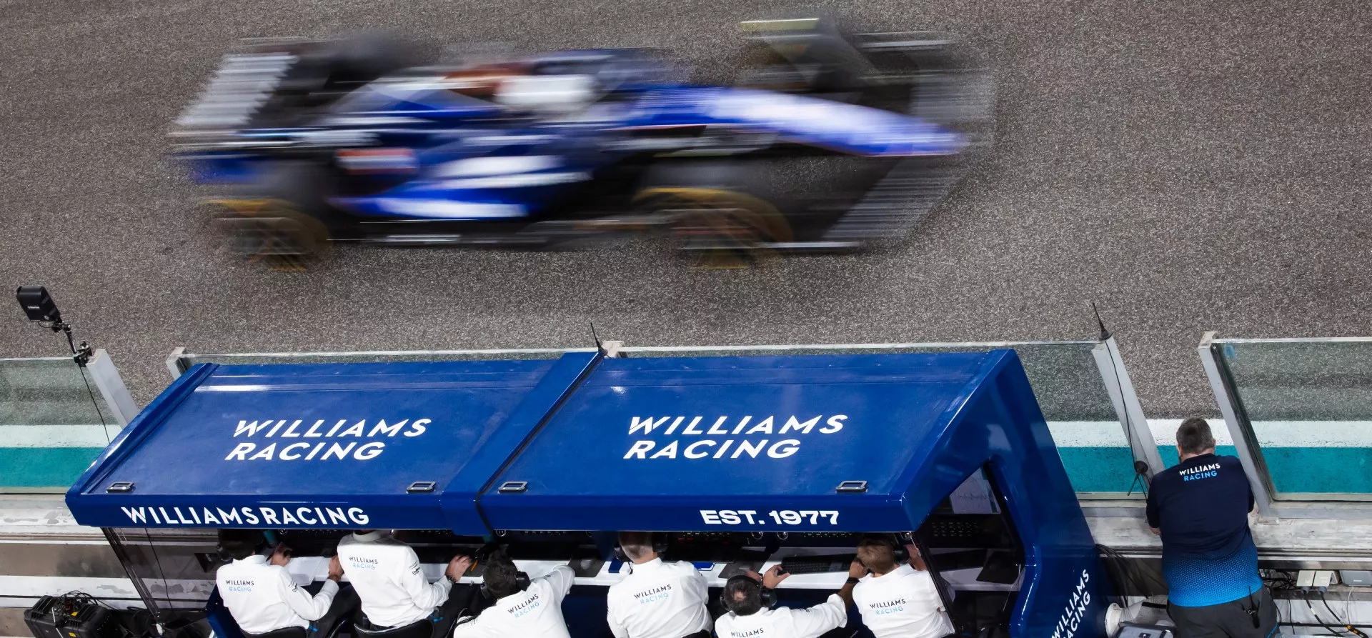 Blue F1 car, blurry, going at high speed past the Williams Racing pitwall